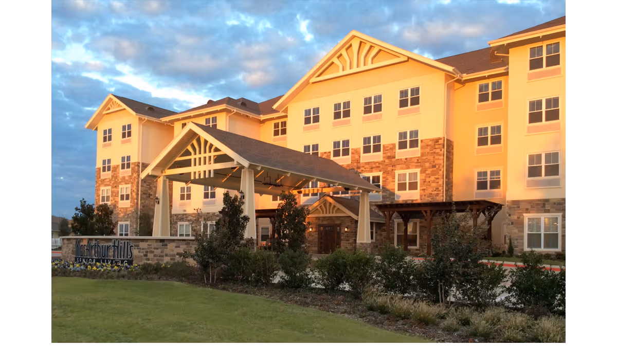 Exterior view of a multi-story senior living facility building named MacArthur Hills, with a covered entrance, stone and beige facade, surrounded by landscaping and greenery under a partly cloudy sky.