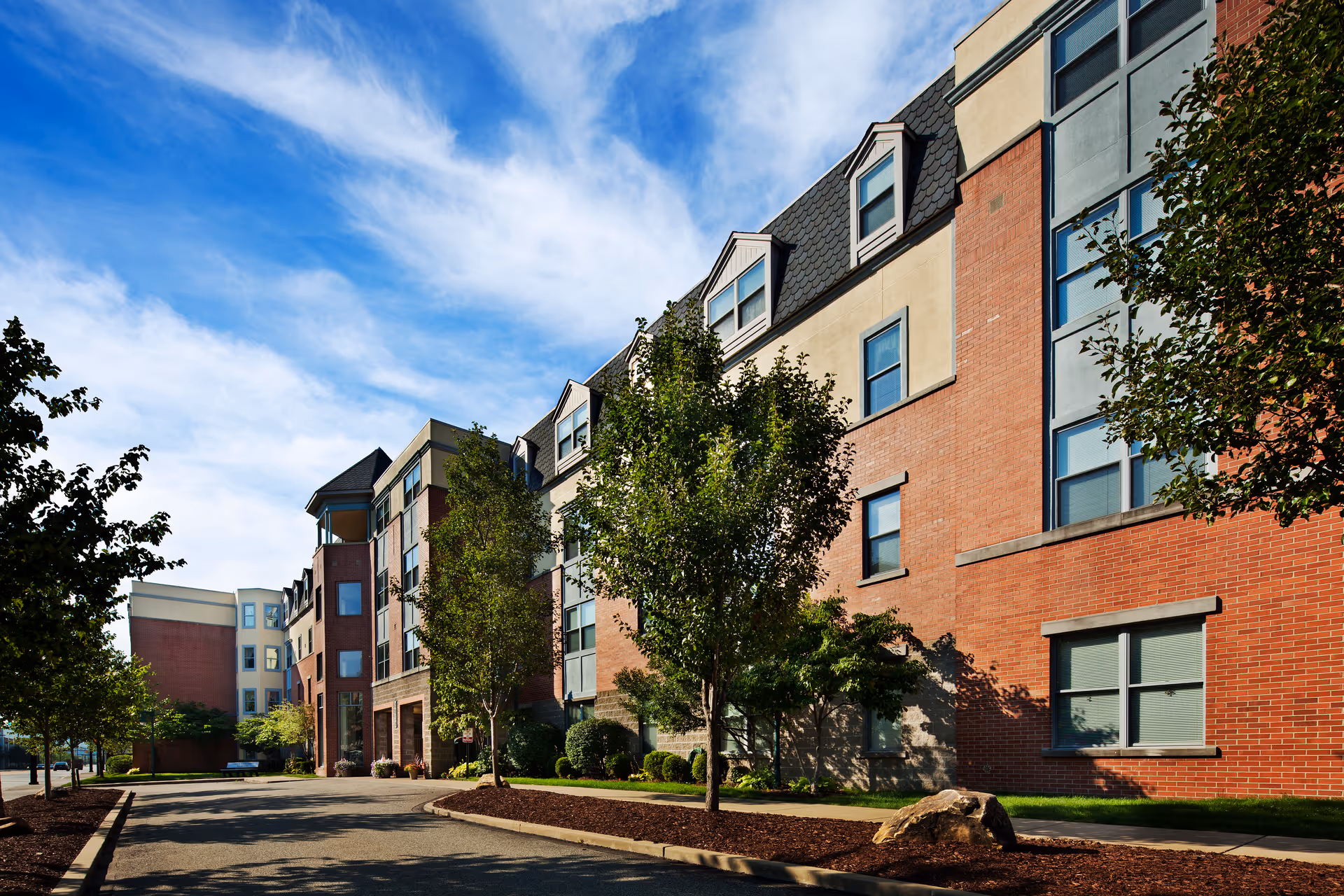 Front exterior of a multi-story brick retirement residence with a driveway, landscaped trees, and a blue sky.