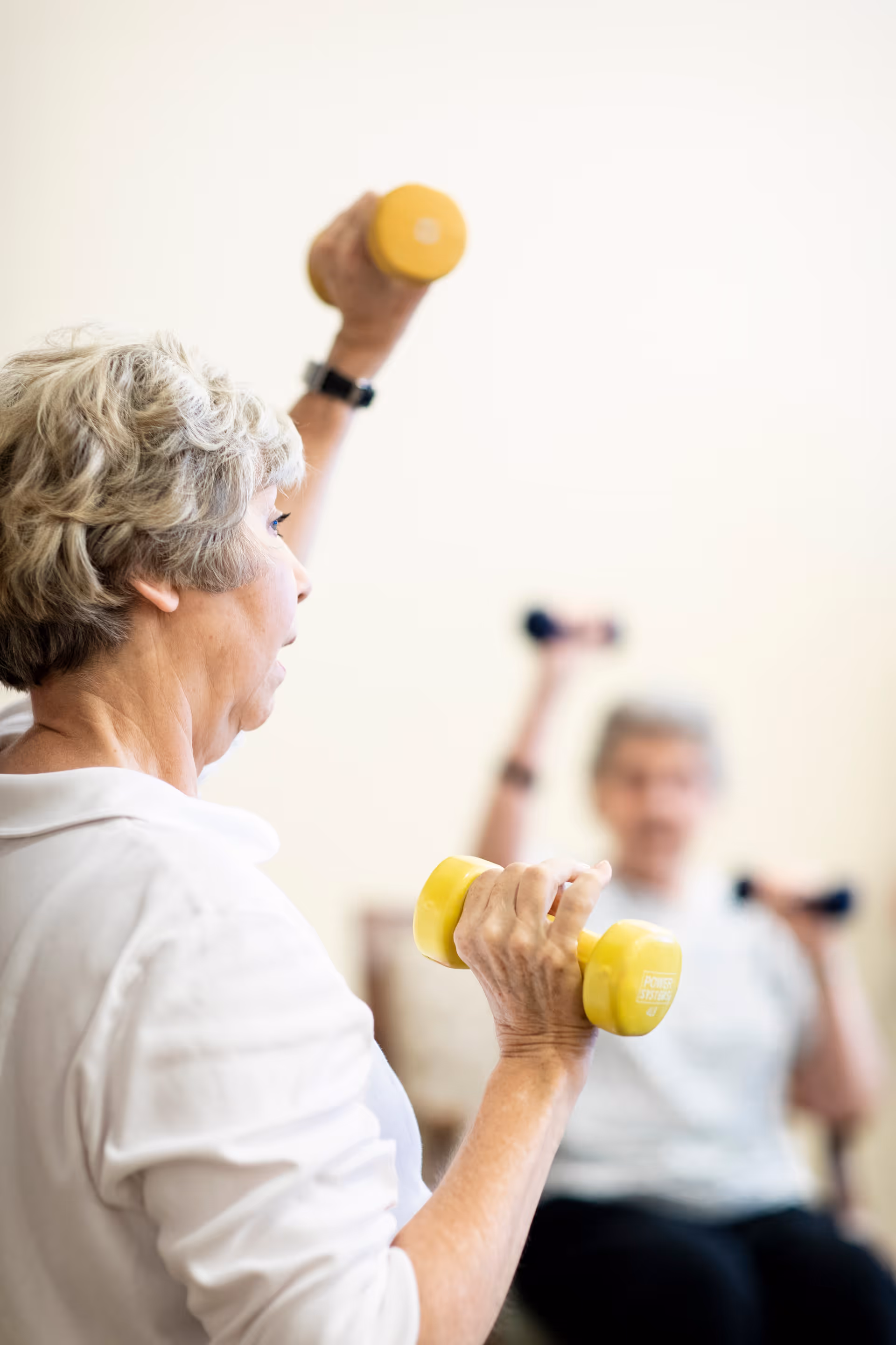 Two elderly women exercising indoors while seated, each holding a pair of small dumbbells and lifting one arm overhead.
