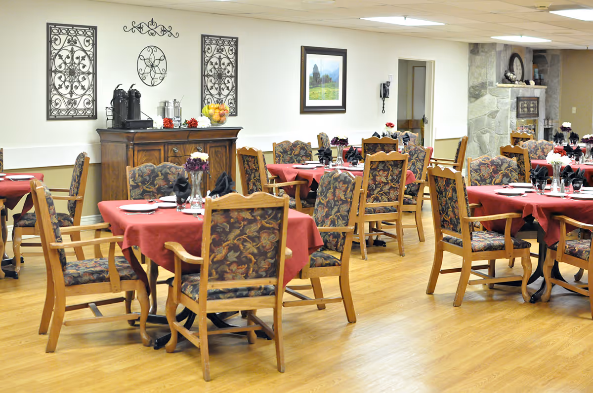 A dining room with multiple wooden tables covered with red tablecloths and surrounded by wooden chairs with floral upholstery. The tables are set with plates, glasses, and black napkins. The room has wooden flooring, a stone fireplace, decorative wall art, and a wooden sideboard with coffee urns and a bowl of fruit.