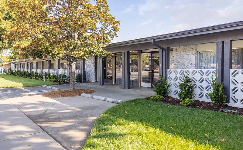 Exterior view of a single-story building with a covered entrance, large windows, and decorative white concrete blocks. There is a tree and well-maintained green grass in front of the building, along with a concrete walkway and parking stops.