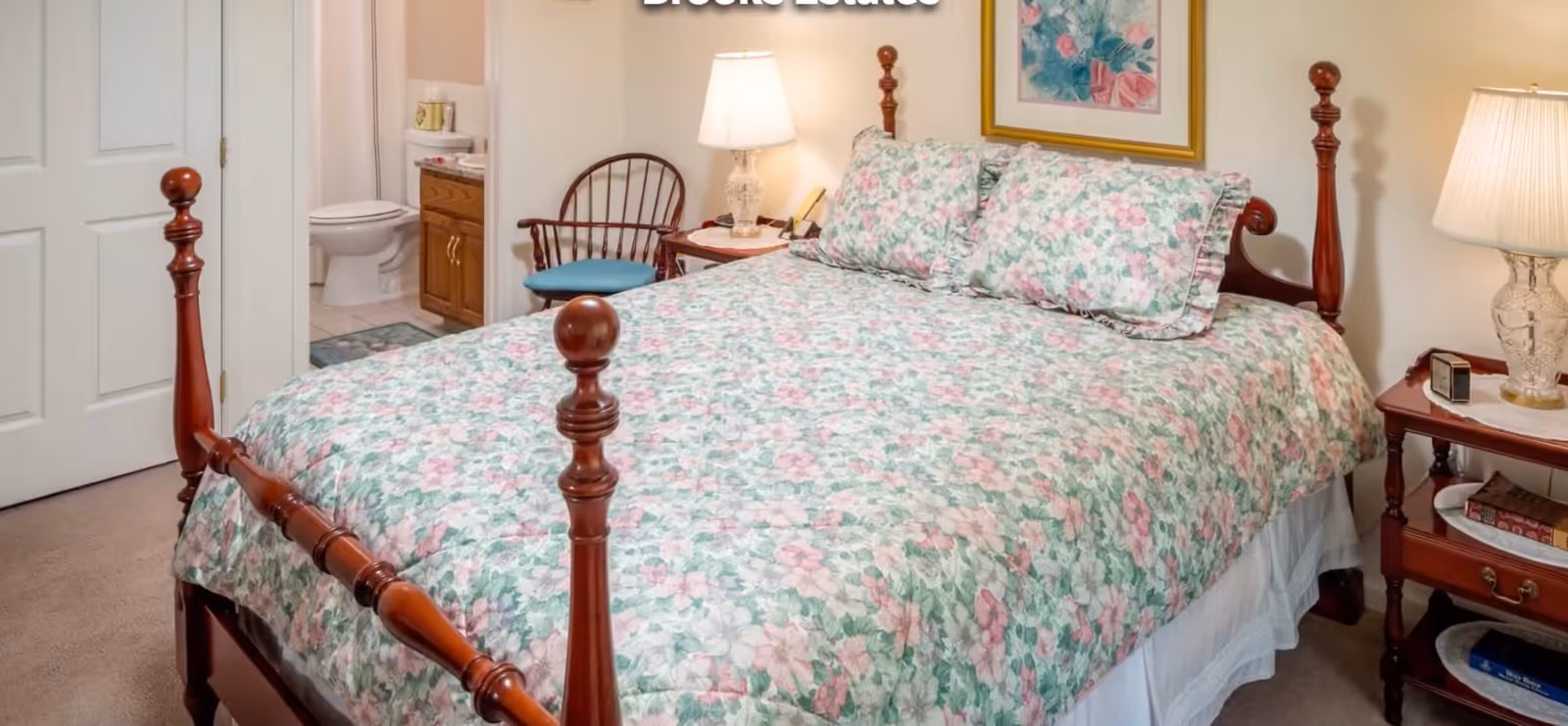 A bedroom with a wooden four-poster bed covered with a floral bedspread and matching pillows. There is a wooden side table with a lamp and books on it, a wooden chair with a blue cushion, and a bathroom visible through an open door showing a toilet and a vanity with a sink.