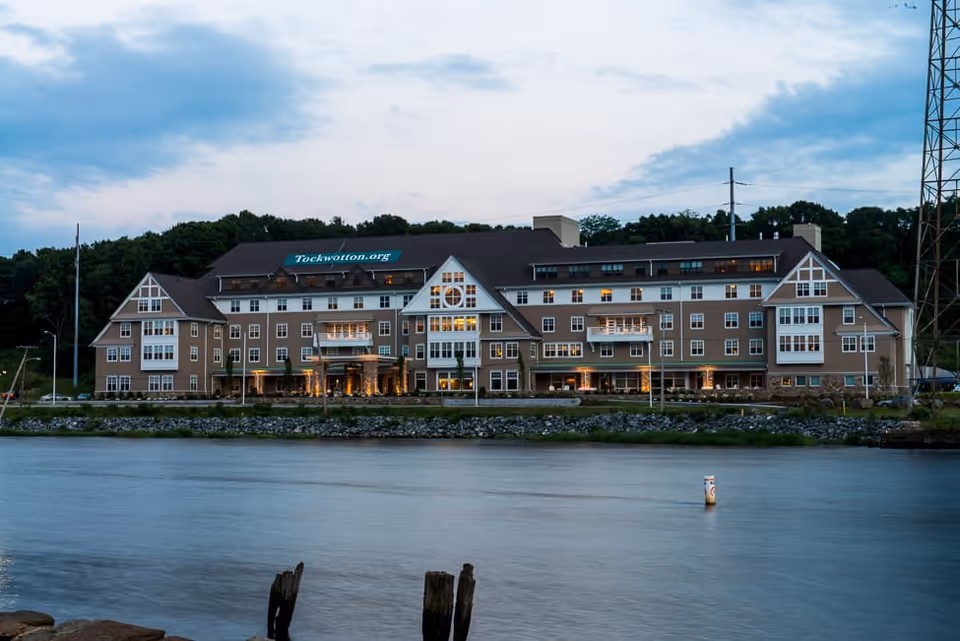 A large multi-story building situated on the waterfront during dusk, with lights glowing from the windows. The building has a brown exterior with white trim and multiple balconies. Trees and a cloudy sky are visible in the background, and calm water is in the foreground.