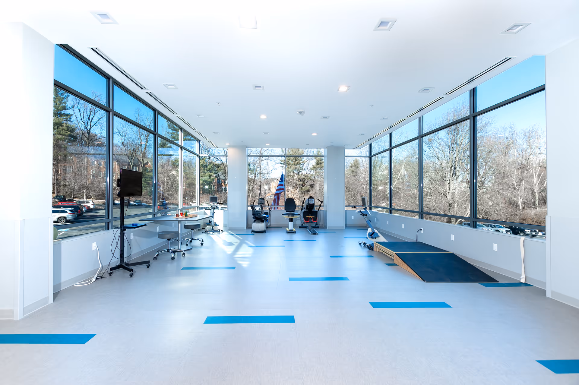 Bright rehabilitation room with large floor-to-ceiling windows on two sides, exercise equipment including stationary bikes and a ramp, and a few chairs and tables along the walls. Outside, trees and parked cars are visible.