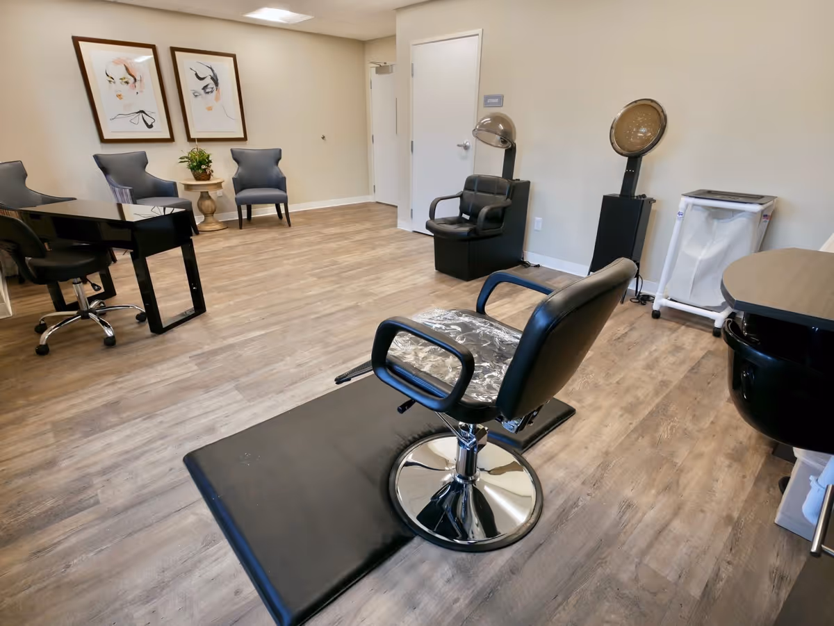 Interior view of a salon area in a senior living facility with a black salon chair on a black mat, two vintage hair dryers, a black sink, three gray chairs against the wall, a small round table with a plant, and two framed abstract face artworks on the wall.