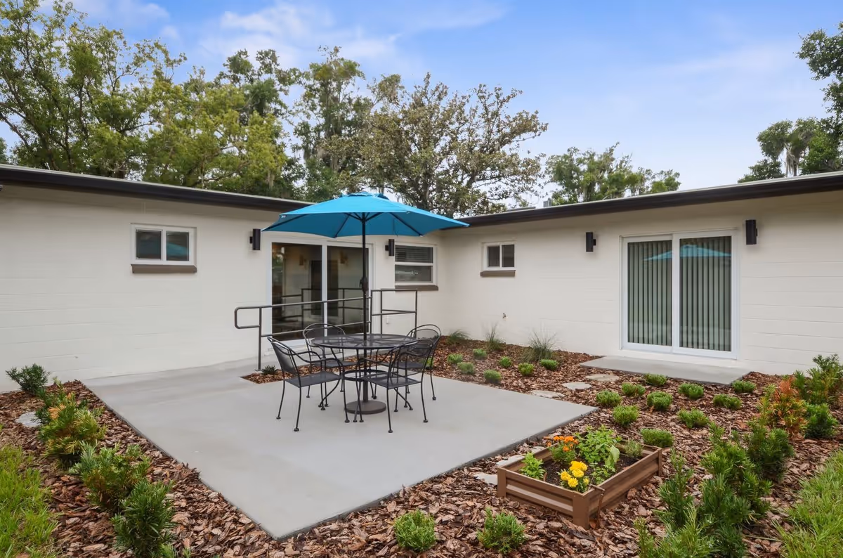 Outdoor patio area at Vivant Senior Living, Memory Care Homes featuring a concrete seating area with a black metal table and four chairs under a blue umbrella. The patio is surrounded by landscaped garden beds with small shrubs and a raised planter box with flowers. The building exterior is white with sliding glass doors and windows, and trees are visible in the background under a partly cloudy sky.