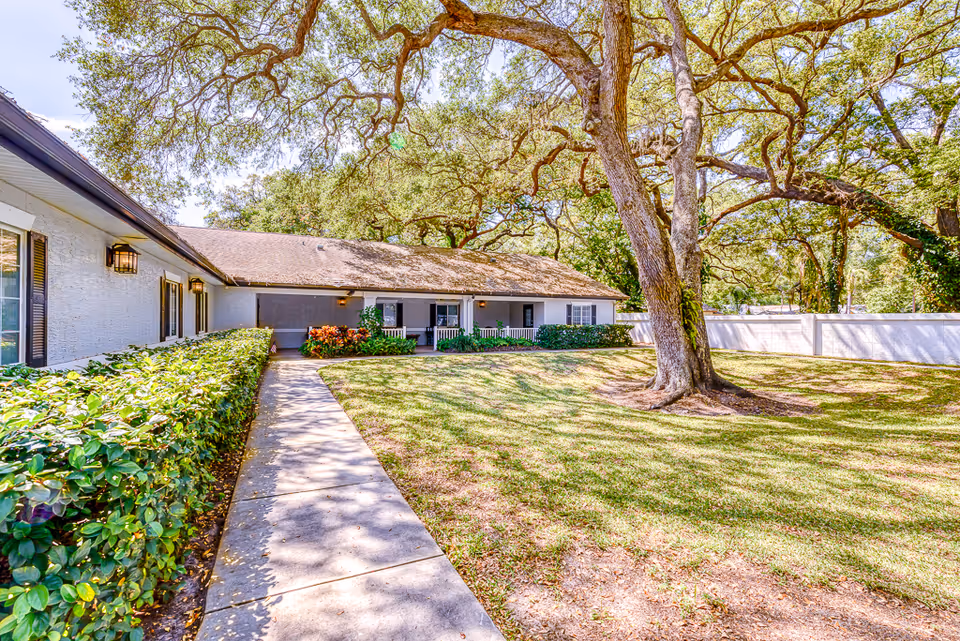 Single-story residential building and courtyard with a large oak tree, walkway, hedges, and lawn.