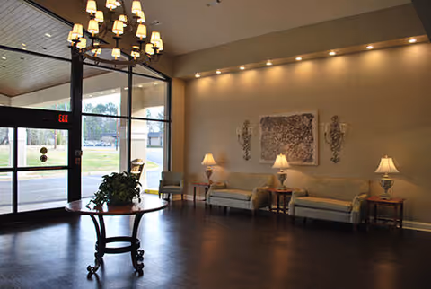 A spacious lobby area with large glass entrance doors, a round wooden table with a plant in the center, two sofas with cushions along the wall, three table lamps, wall sconces, and a chandelier hanging from the ceiling.