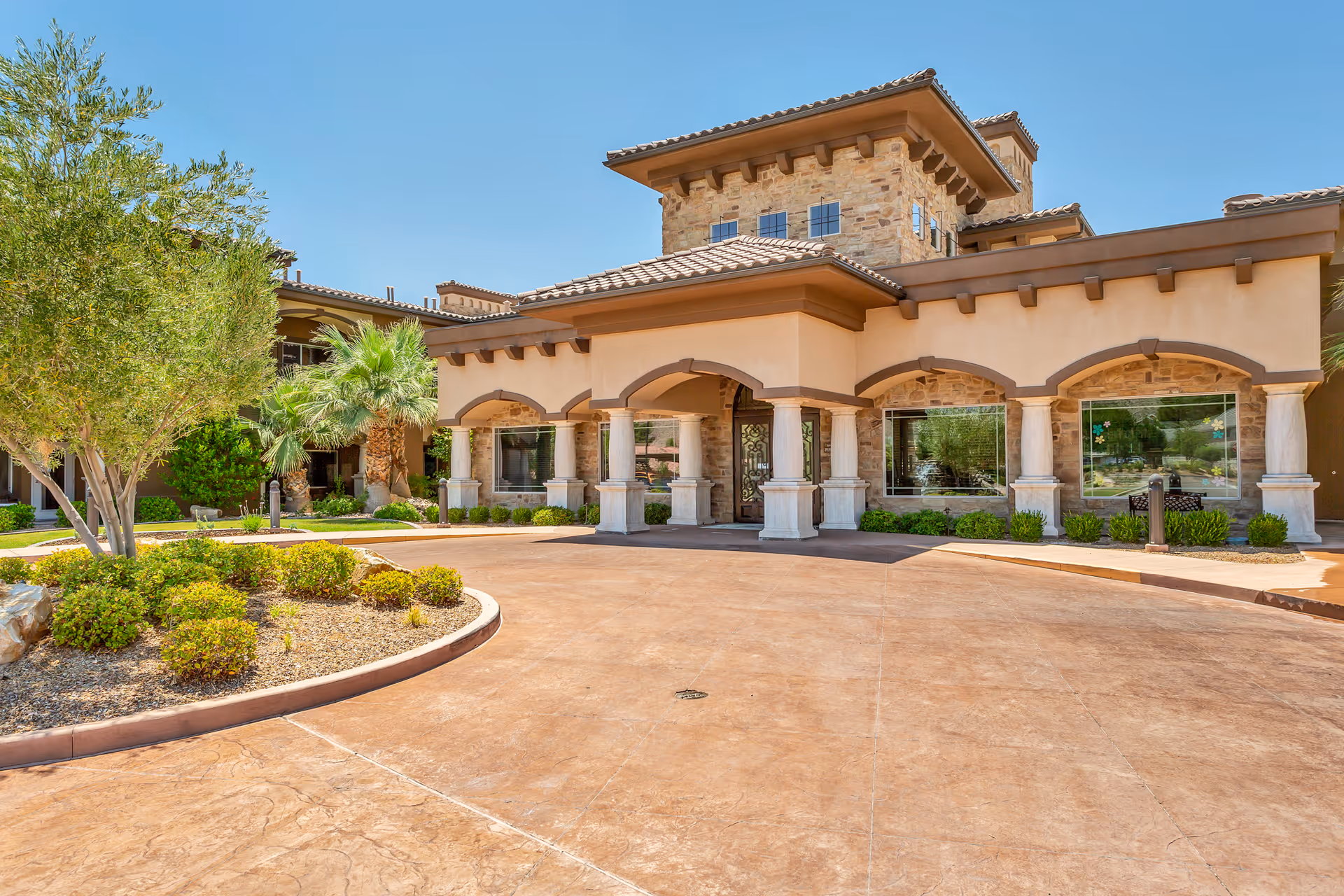 Front entrance of a Mediterranean-style senior living building with arched columns, large windows, and a landscaped circular driveway.