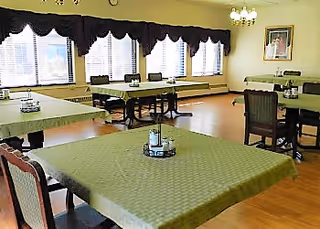 Bright dining room with several tables covered in green tablecloths and wooden chairs arranged under large windows.