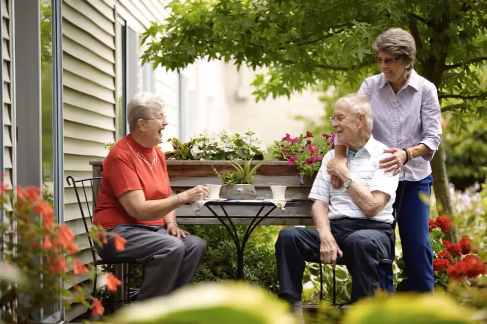 Three elderly people enjoying a conversation outdoors in a garden area. One woman in a red shirt is sitting on a chair holding a cup, an elderly man in a striped shirt is seated next to her, and another woman stands behind the man with her hands on his shoulders. They are surrounded by greenery and flowers near a building with siding.