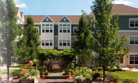 Exterior view of a senior living facility with a well-maintained garden area featuring trees, shrubs, and benches in front of a multi-story building with multiple windows and a brown roof under a blue sky with some clouds.