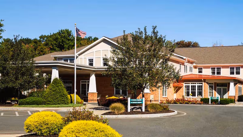 Front entrance of a brick senior living facility with a circular driveway, landscaped shrubs, and an American flag.