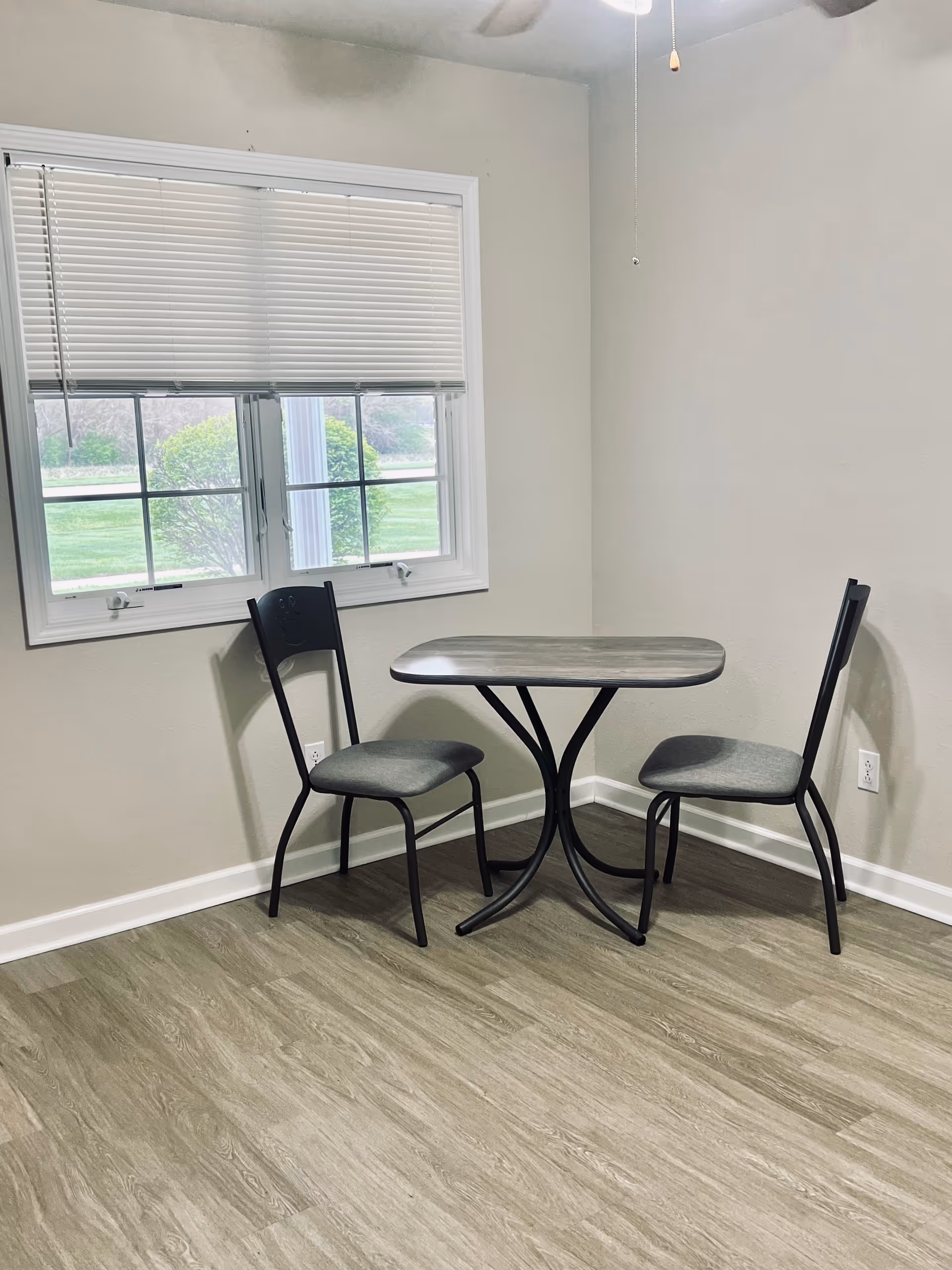 A small dining area with a rectangular table and two chairs with gray cushions. The table and chairs have black metal frames. There is a window with white blinds partially open, showing greenery outside. The room has light-colored walls and wood-patterned flooring.