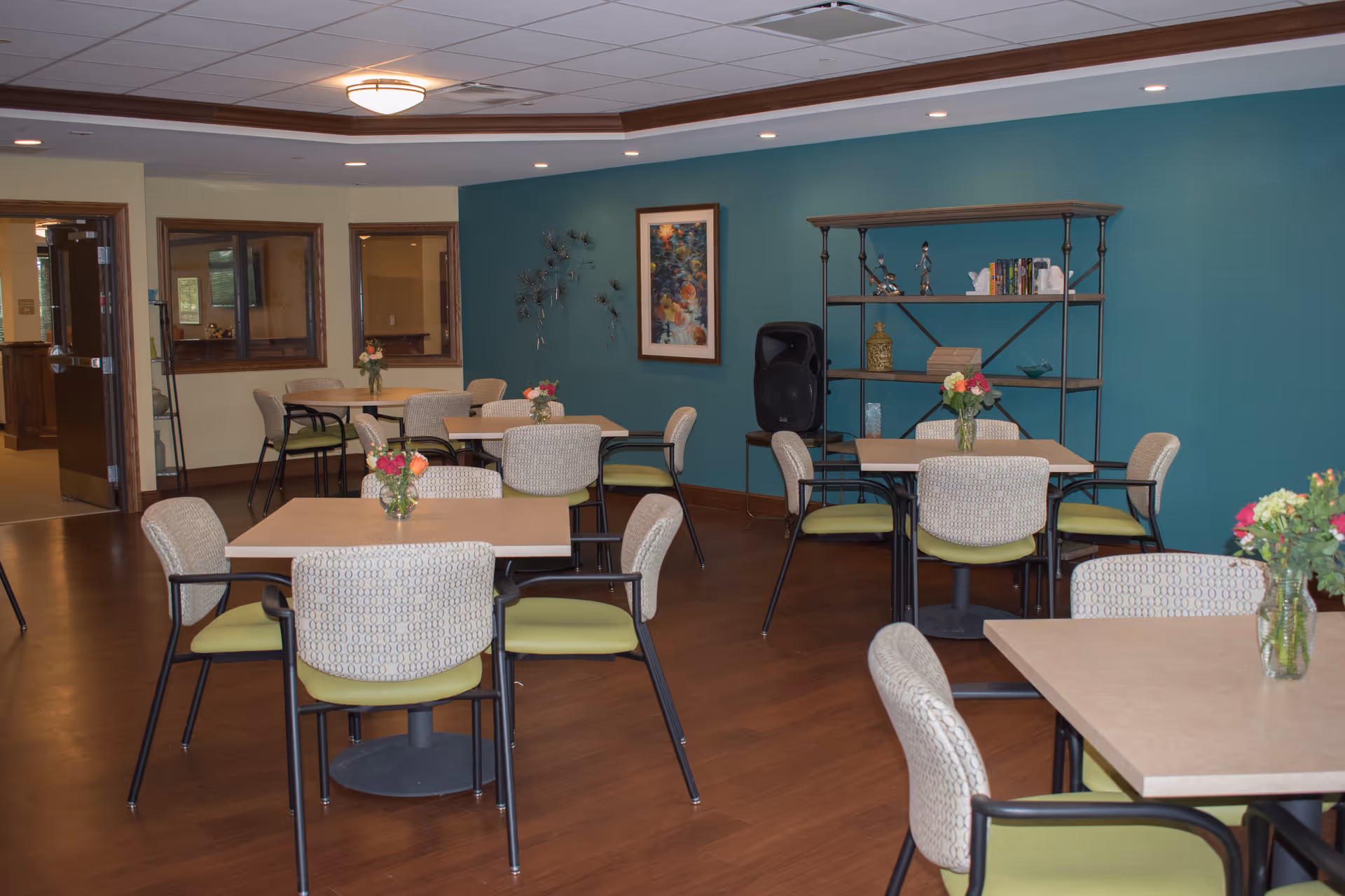 Dining/activity room with multiple tables and chairs, floral centerpieces, and a teal accent wall with shelving and artwork.