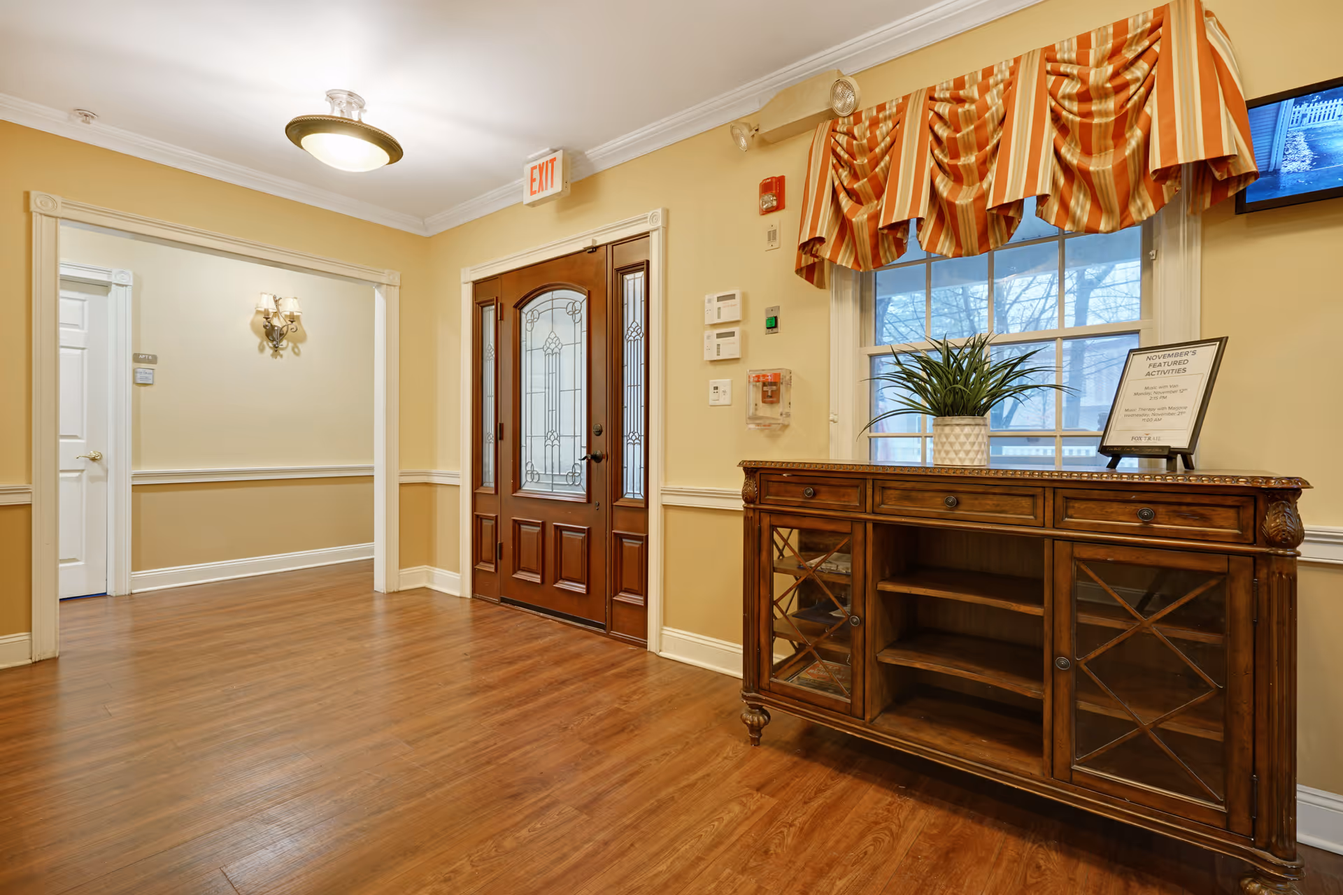 Interior view of a hallway in a senior living facility with wooden flooring, beige walls, a wooden door with glass panels, a wooden cabinet with glass doors, a potted plant, a framed sign, and a window with striped valance curtains.
