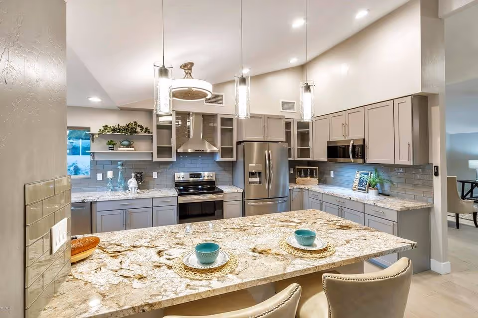 Modern kitchen with granite countertops, gray cabinets, stainless steel appliances including a refrigerator, oven, and microwave. Three pendant lights hang above the kitchen island, which has two place settings with turquoise bowls and plates. The kitchen has a light gray tile backsplash and an open layout leading to a dining area.