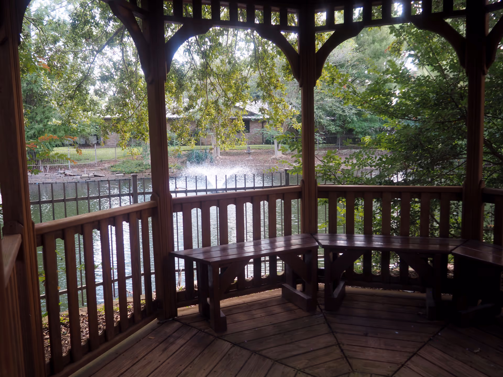 View from inside a wooden gazebo overlooking a pond with a water fountain, surrounded by trees and greenery, with a building visible in the background.