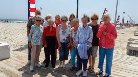 A group of elderly people standing together on a wooden boardwalk near a sandy beach with American flags on poles lining the walkway. The ocean is visible in the background under a clear sky.