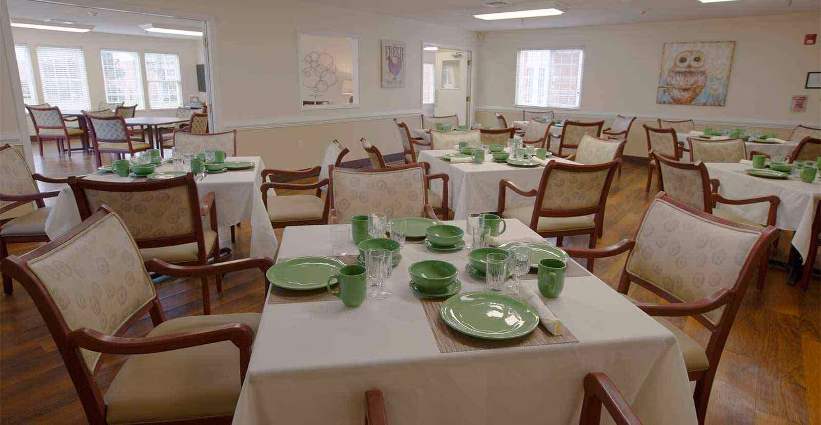 A dining room in a senior living facility with multiple tables covered in white tablecloths. Each table is set with green plates, bowls, mugs, and clear drinking glasses. The room has wooden floors, beige walls, and several windows letting in natural light. There is a painting of an owl on one wall and comfortable wooden chairs with cushioned seats around the tables.