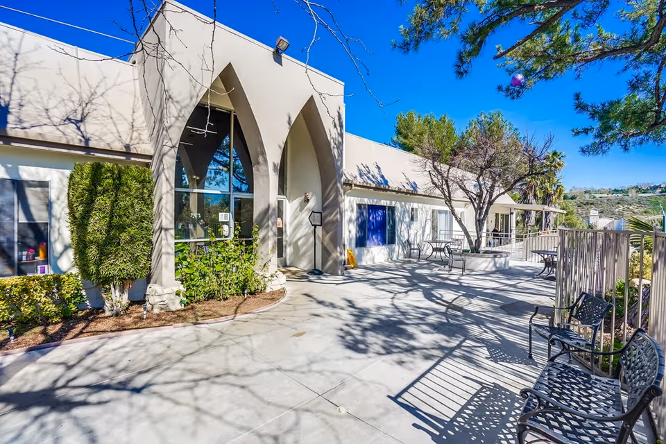 Outdoor entrance and patio of a light-colored senior living facility with an arched doorway, benches, tables, trees, and a clear blue sky.