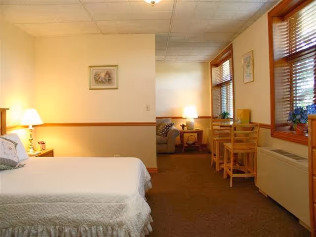 Cozy senior living studio with a bed on the left, a small seating area in the back, and a table with chairs by windows on the right.