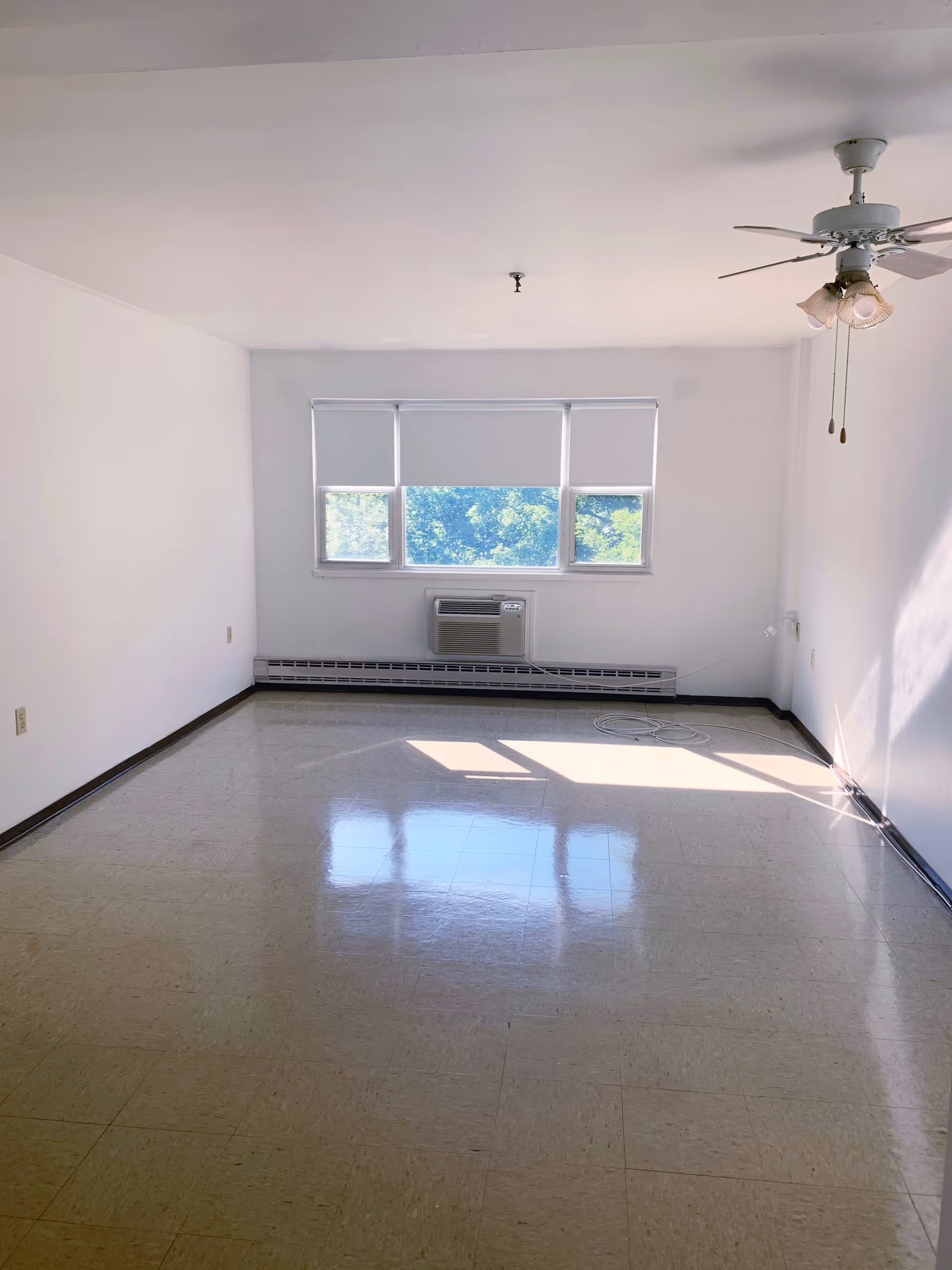 Empty room with white walls, a ceiling fan with lights, a window with three sections partially covered by blinds, an air conditioning unit below the window, and a shiny tiled floor reflecting the window light.