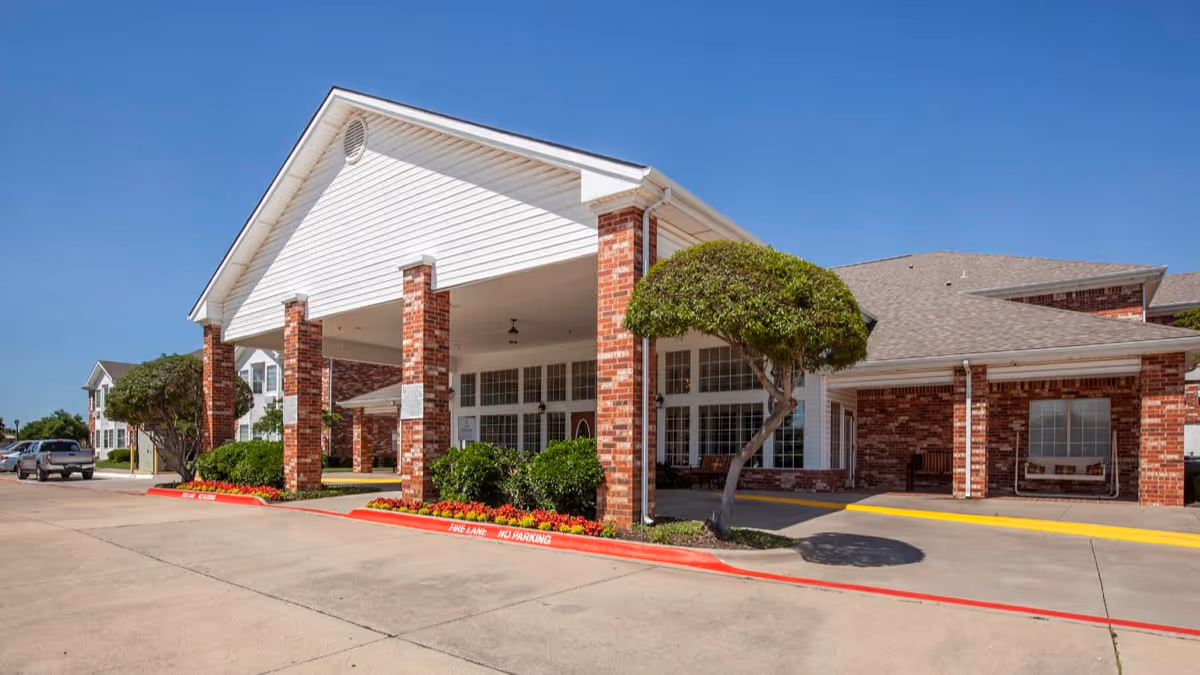 Exterior view of a senior living facility building with a covered entrance supported by brick columns, neatly trimmed bushes, a small tree, and a clear blue sky.
