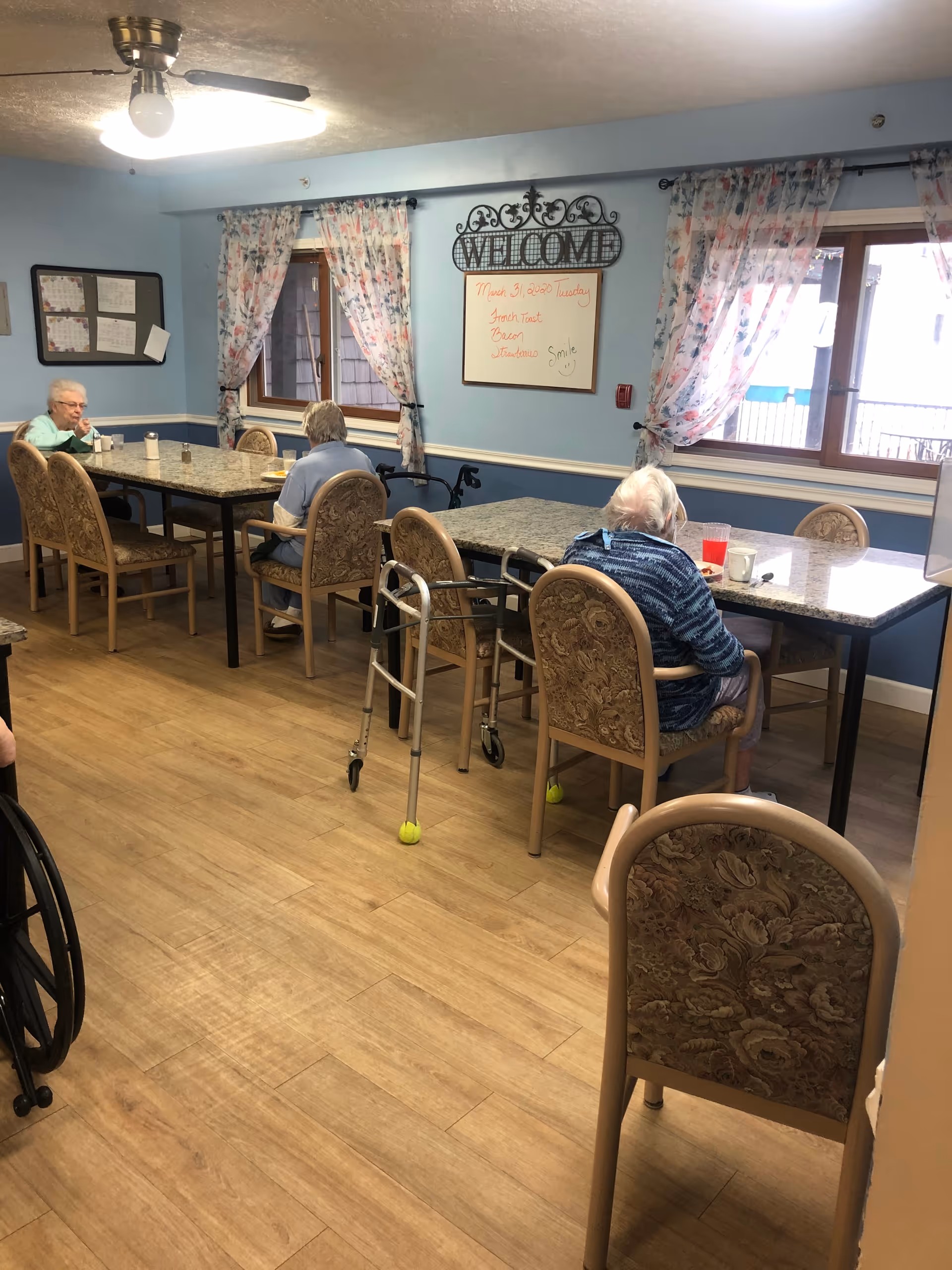 A dining room in an assisted living facility with elderly residents seated at tables. The room has light blue walls, floral curtains on the windows, and a wooden floor. There is a whiteboard on the wall with a welcome sign above it, and mobility aids such as walkers and a wheelchair are visible.