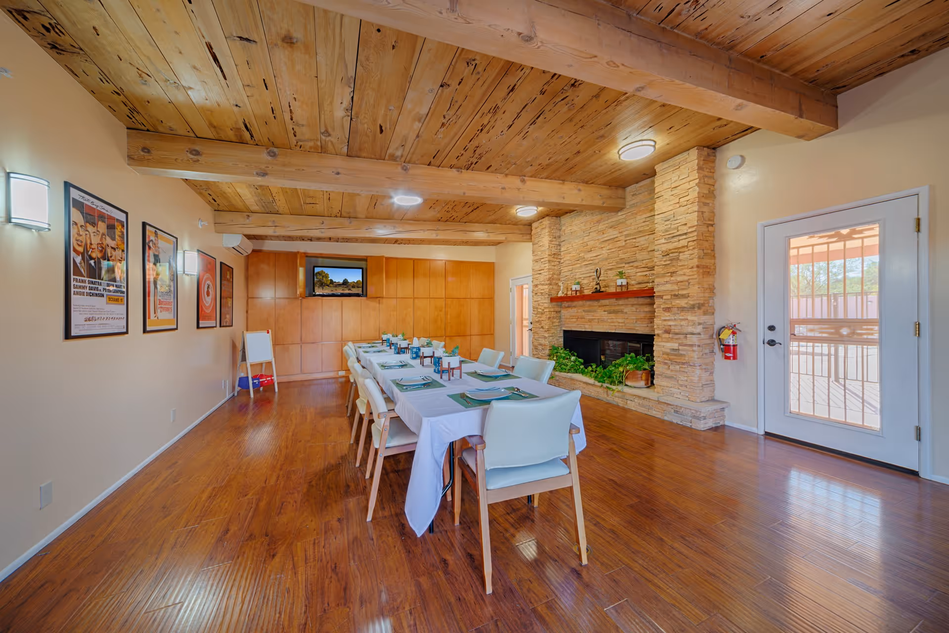 A dining room with a long table set for a meal, surrounded by chairs. The room features a wooden ceiling with exposed beams, a stone fireplace with plants on the hearth, wooden flooring, and a door with a window leading outside. There are framed posters on one wall and a television mounted on the far wall.