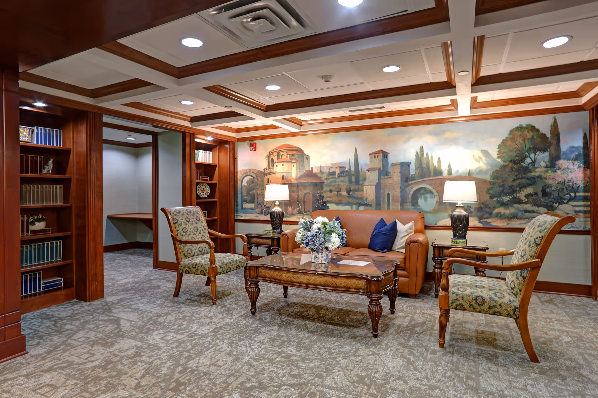 A cozy sitting area with a brown leather sofa, two patterned armchairs, a wooden coffee table with a floral arrangement, and two table lamps. The room features wood trim, built-in bookshelves with books and decorative items, and a large mural on the wall depicting a scenic landscape with buildings, trees, and a bridge.