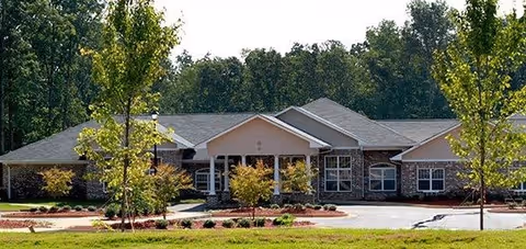 Single-story building with a gabled roof surrounded by trees and landscaping, featuring a driveway and entrance area in front.