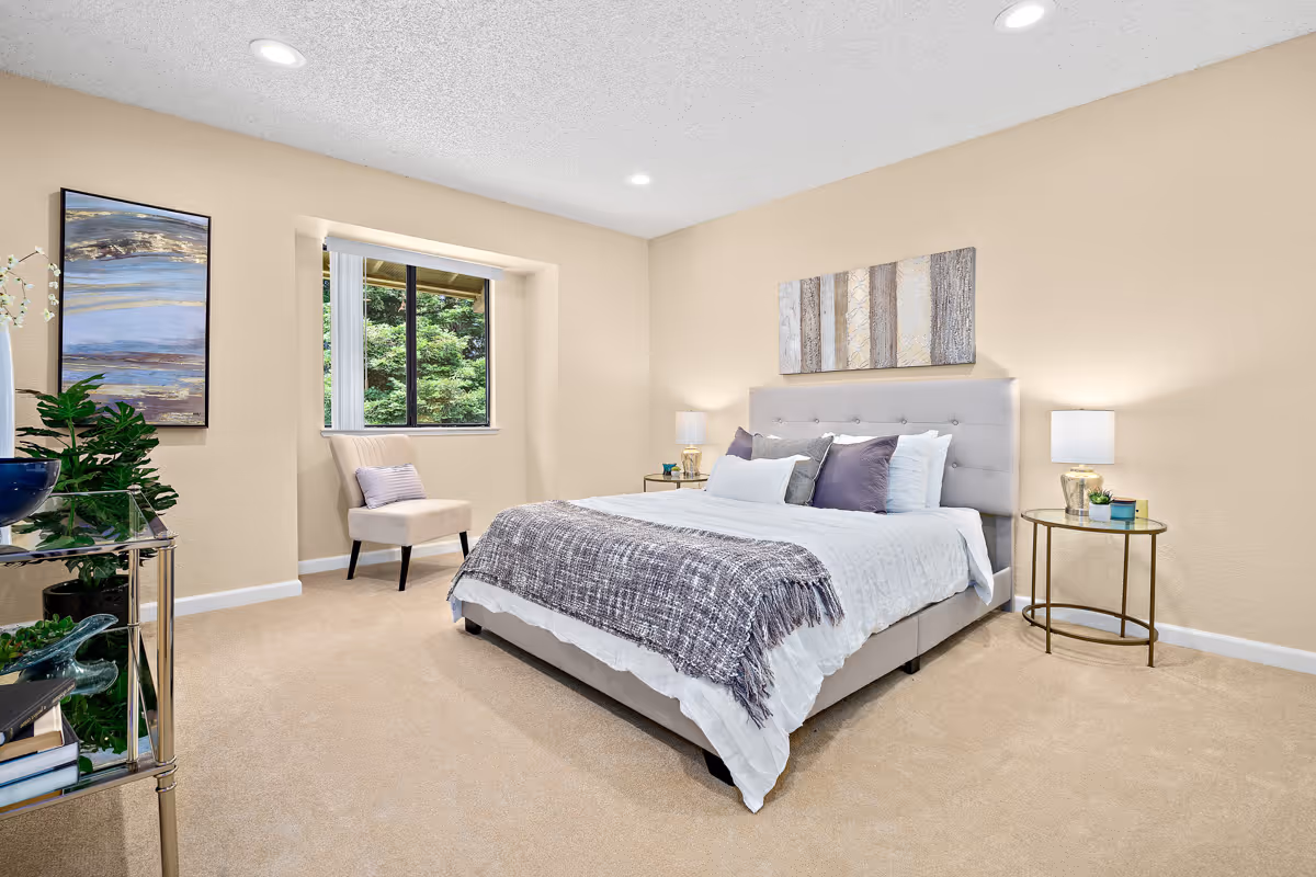 A bright and cozy bedroom with a large bed featuring white and gray bedding and multiple pillows. There are two round bedside tables with lamps on each side of the bed. A beige chair is placed near a window with a view of greenery outside. The walls are painted light beige, and there is a piece of abstract artwork hanging above the bed. A glass shelf with plants and decorative items is visible on the left side of the room.