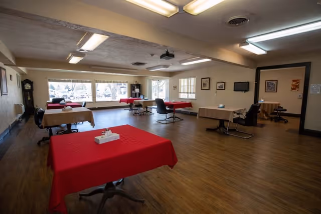 A spacious room with several tables covered in red and beige tablecloths, each with a tissue box on top. The room has wooden flooring, large windows letting in natural light, a grandfather clock, framed pictures on the walls, and office chairs around the tables.