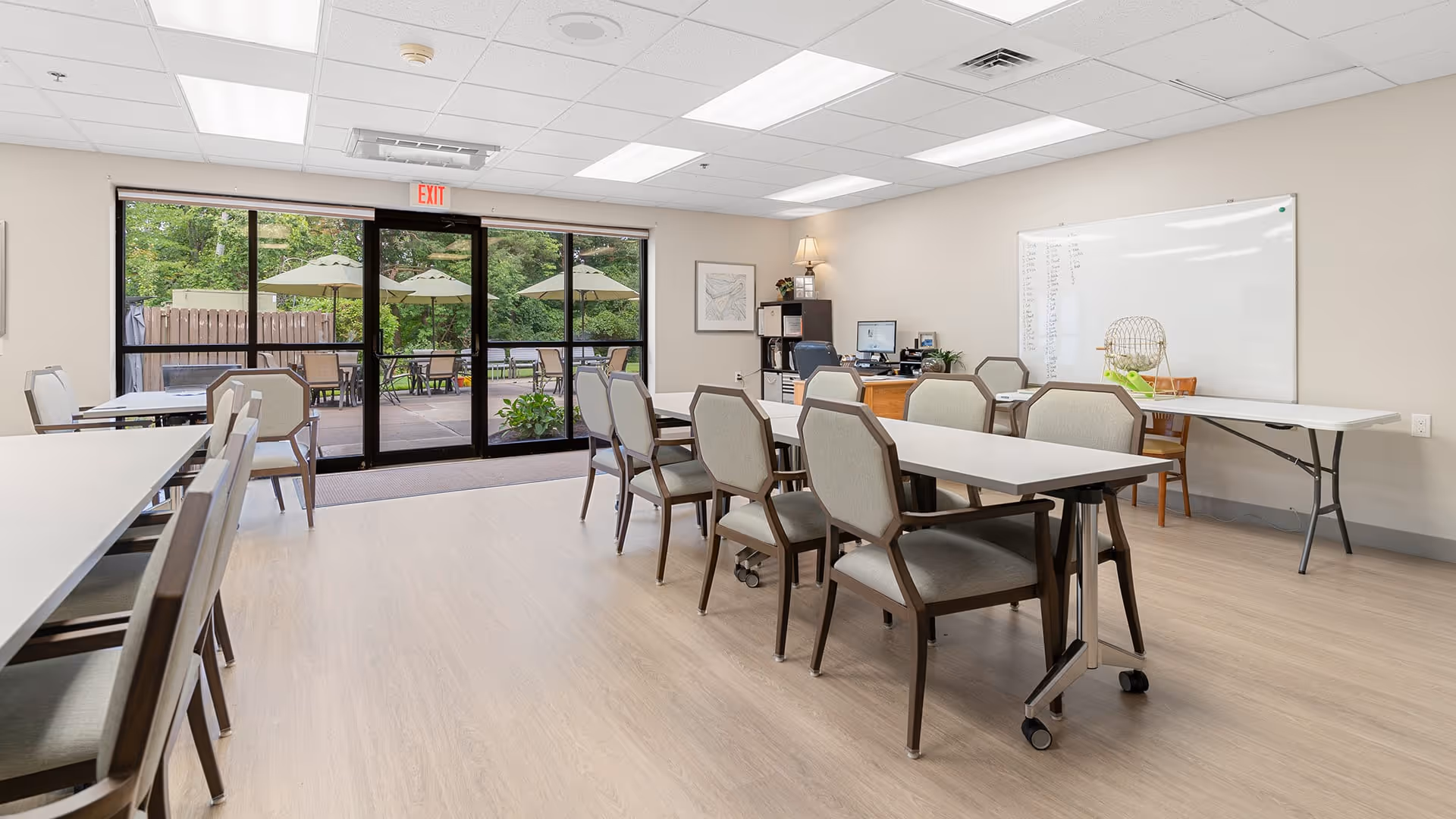 A bright room with several tables and chairs arranged for group seating. There is a large whiteboard on the wall with some writing on it, a desk with a computer and lamp in the corner, and large glass doors leading to an outdoor patio area with umbrellas and chairs. The room has light-colored walls and flooring.