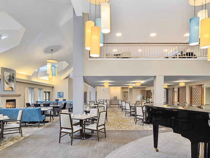 Spacious, well-lit senior living dining and lounge area with tables and chairs, a grand piano, and a mezzanine level.