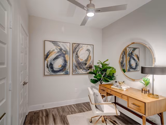 A modern home office with a wooden desk, a white swivel chair, a round mirror on the wall, two abstract paintings, a potted plant, and a ceiling fan with a light. The room has light-colored walls and wood flooring.