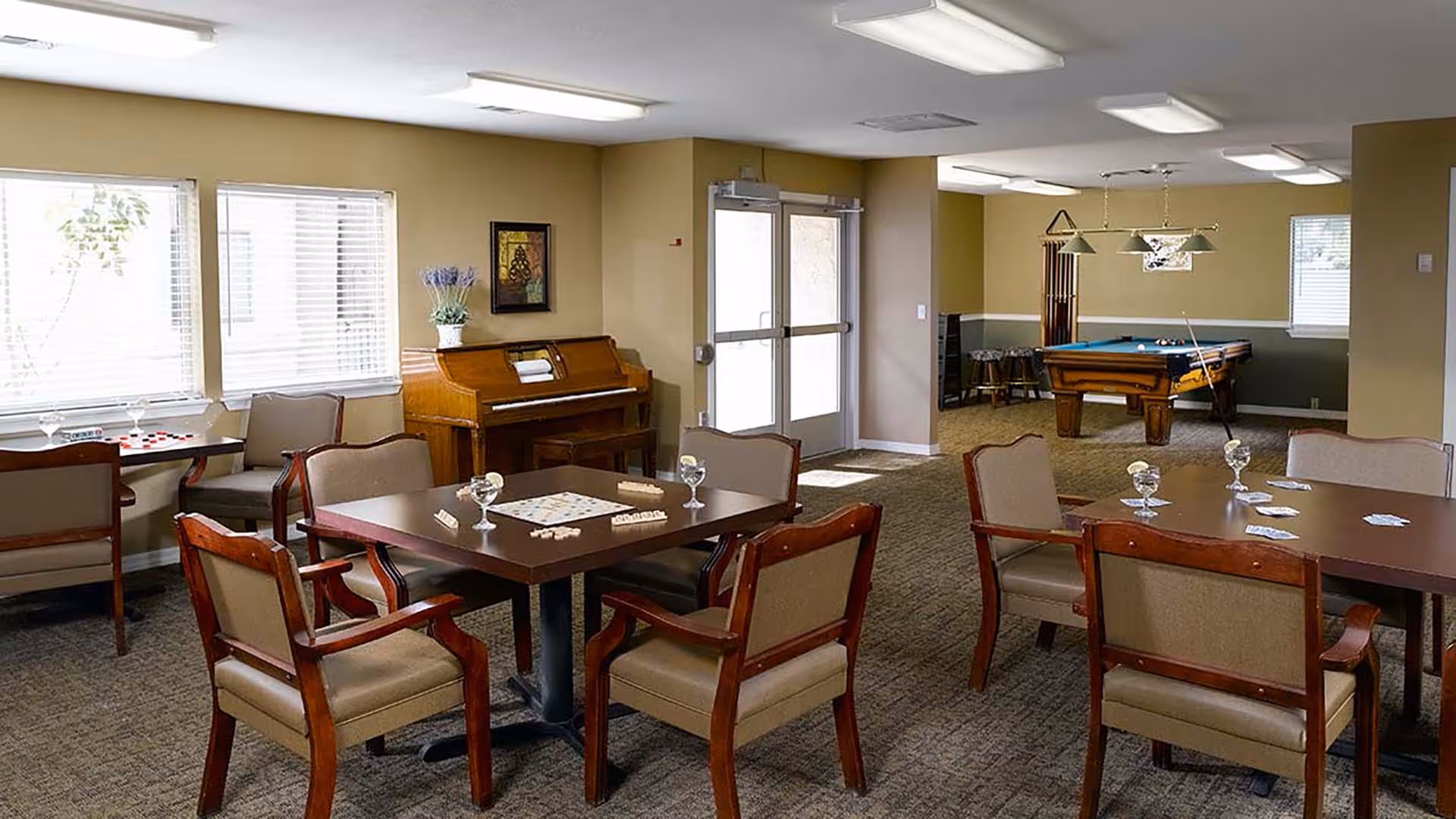 A spacious common room with several tables and chairs arranged for games, including cards and checkers. There is a piano against the wall with a plant on top, and a pool table with hanging lights in the background. The room has beige walls, carpeted floor, and large windows letting in natural light.