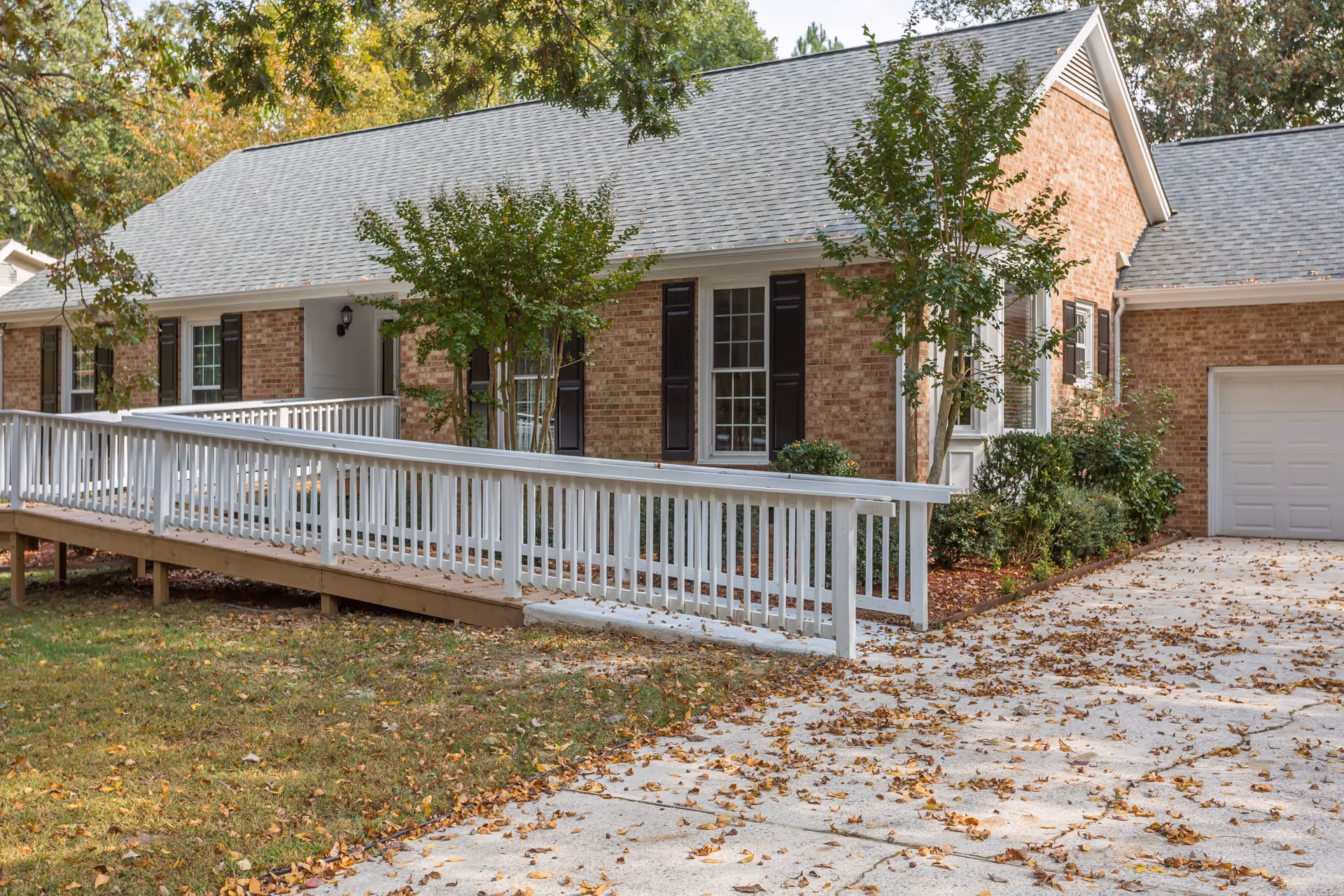 Exterior view of a single-story brick building with a gray shingled roof, black window shutters, and a white wooden ramp leading to the entrance. The surrounding area has trees with green leaves and a driveway covered with fallen autumn leaves.