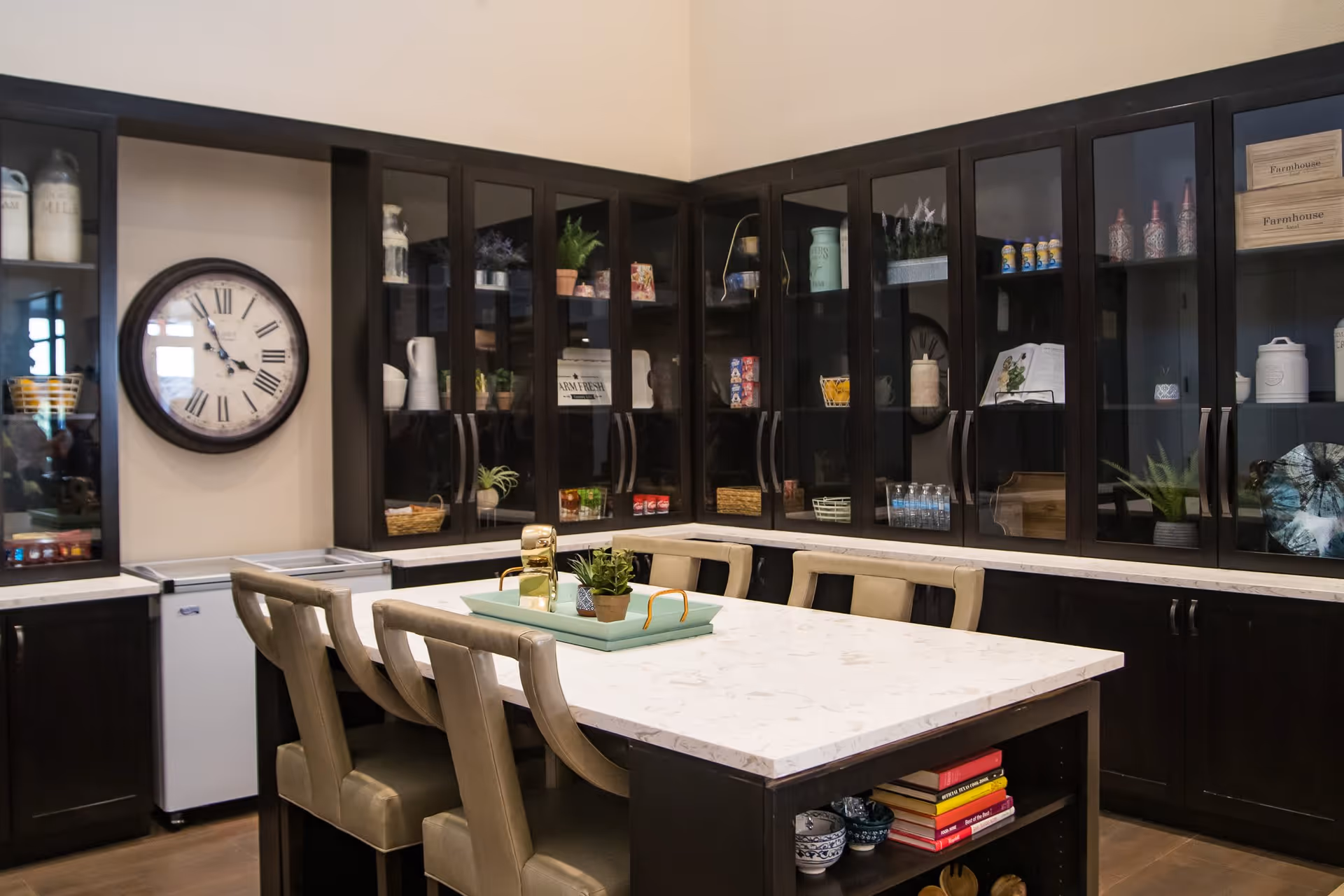 Interior view of a kitchen or dining area with a large marble countertop island surrounded by beige cushioned chairs. The background features dark wood cabinets with glass doors displaying various decorative items, plants, and kitchenware. A large round wall clock is mounted on the wall.