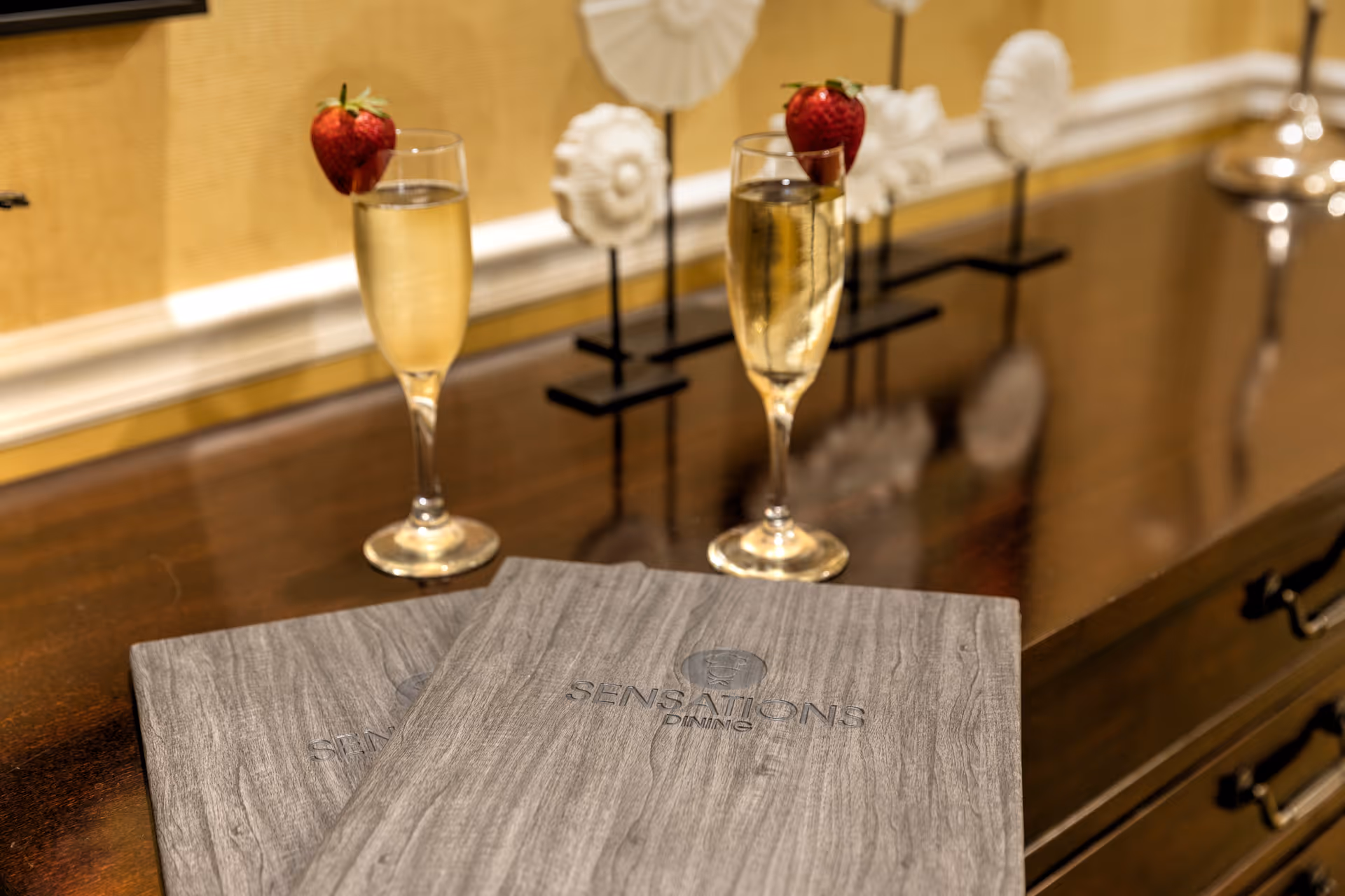 Two glasses of champagne garnished with strawberries placed on a wooden surface next to two closed menus labeled 'Sensations Dining'. Decorative white sculptures are in the background.