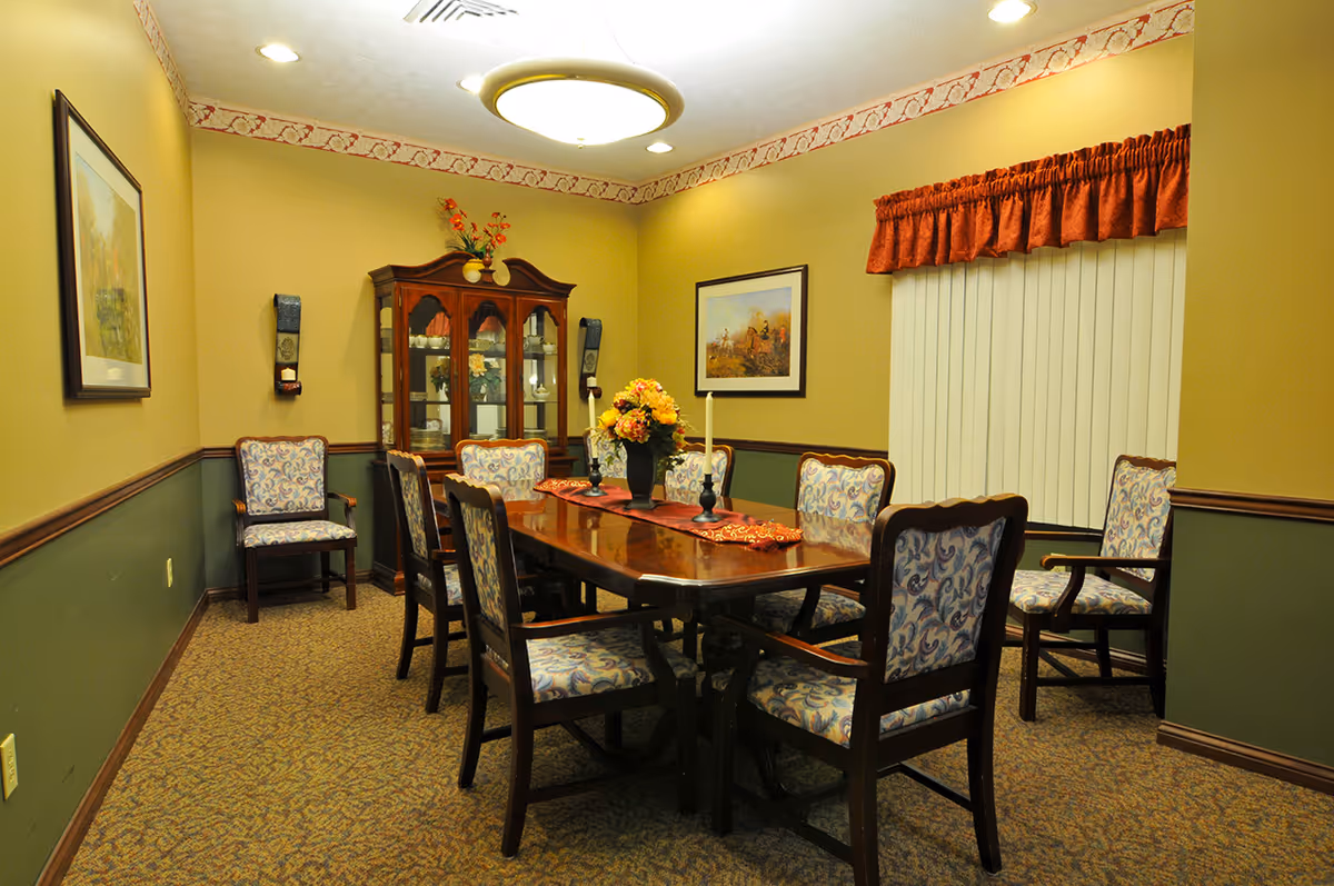 A formal dining room with a polished wooden table surrounded by eight upholstered chairs with patterned fabric. The room has beige and green walls with a decorative border near the ceiling. A wooden china cabinet with glass doors displays dishes and decorative items. Two framed paintings hang on the walls, and a window with vertical blinds and a red valance is visible. A ceiling light fixture illuminates the room.