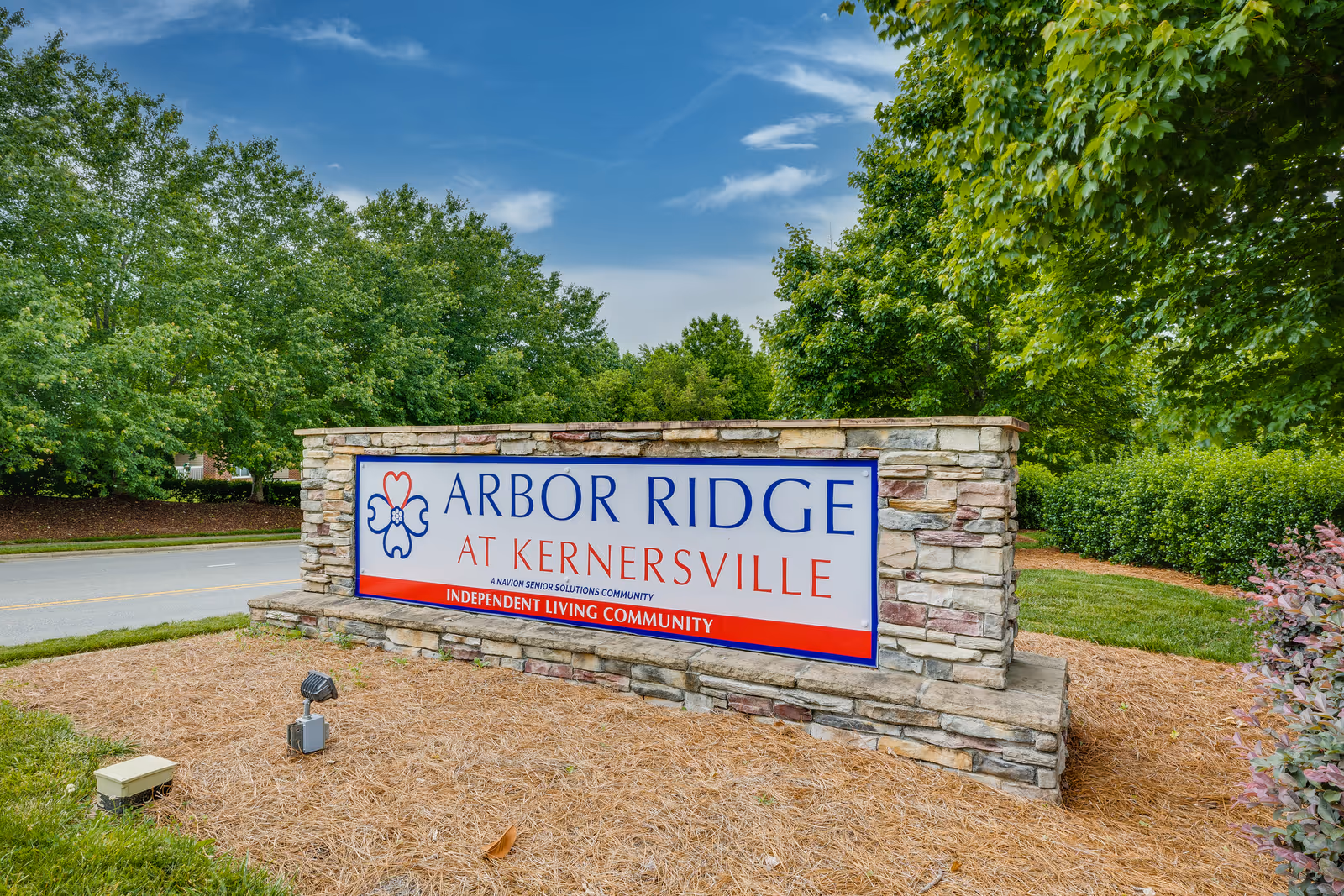 Stone sign for Arbor Ridge at Kernersville, an independent living community, surrounded by greenery and trees with a blue sky background.