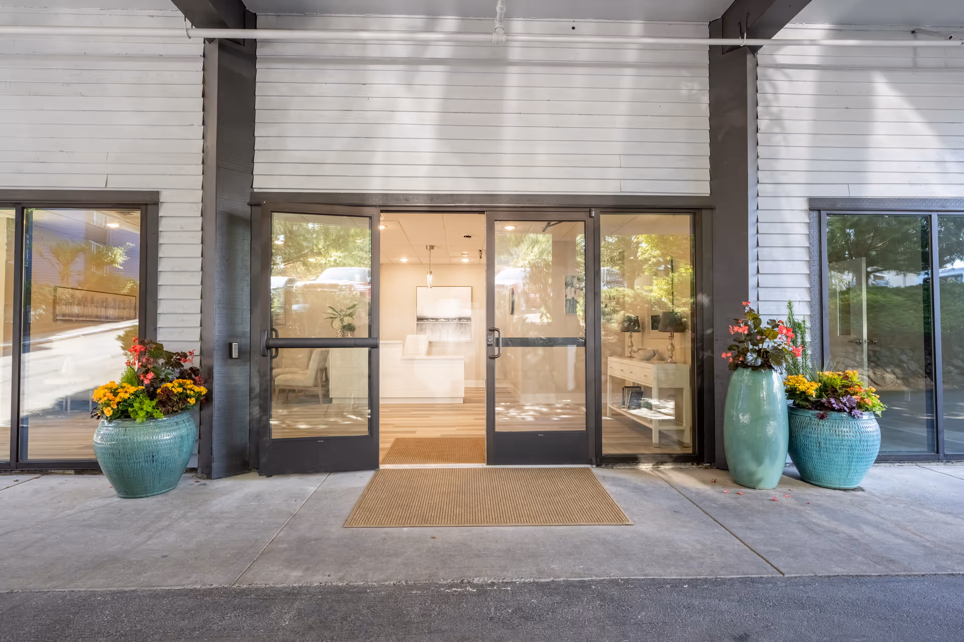 Entrance to a senior living facility with glass double doors, flanked by large green planters filled with colorful flowers. Inside, a reception desk and seating area are visible through the doors.