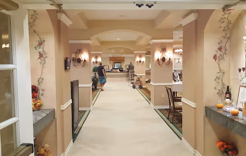 Interior hallway of a senior living facility with beige walls decorated with floral murals and sconces. The hallway leads to a reception or common area where two people are visible, one walking and another standing near a counter. There are chairs and tables on the right side and decorative items on ledges on both sides of the hallway.