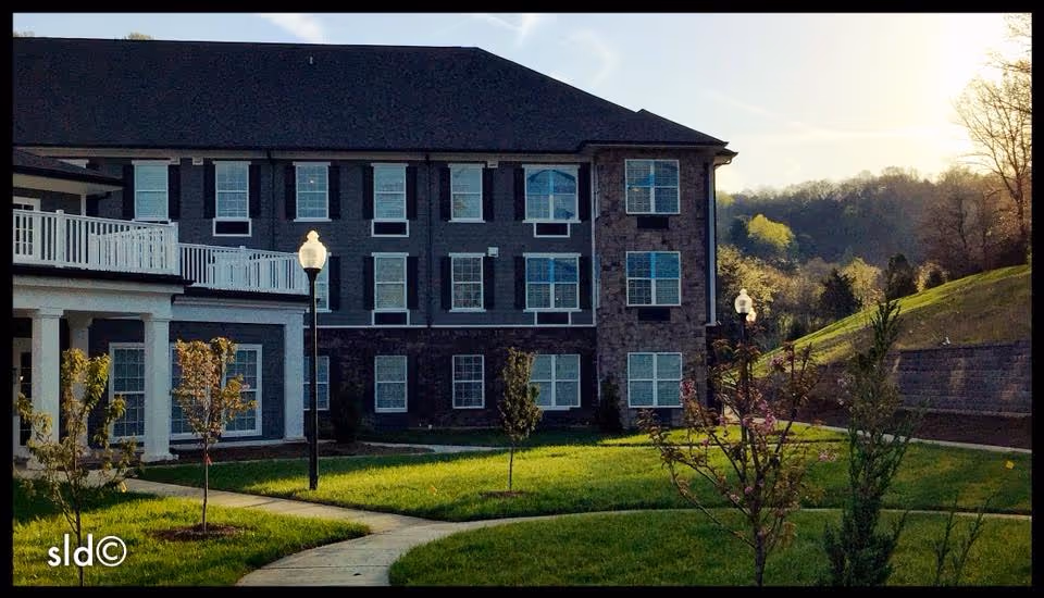 Exterior view of a multi-story senior living facility building with multiple windows and a dark roof. The foreground shows a well-maintained lawn with young trees, a curved concrete pathway, and lampposts. The background includes a wooded hillside under a clear sky with the sun low on the horizon.