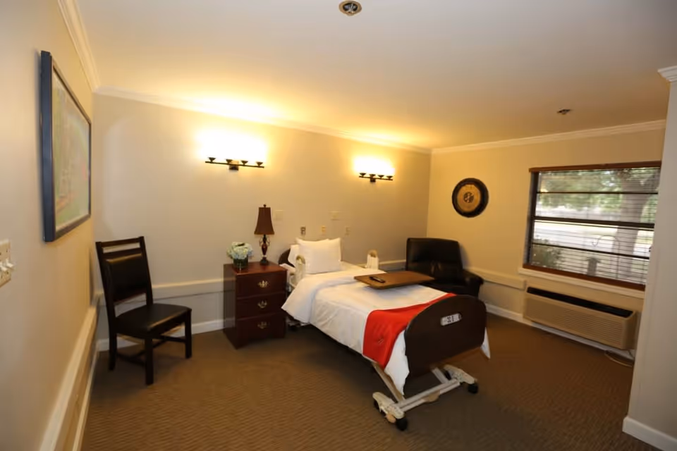 A well-lit assisted living bedroom with a single hospital-style bed covered with white linens and a red blanket. The room includes a wooden nightstand with a lamp and a flower vase, a black chair, a wall clock, a window with blinds, and a heating/cooling unit beneath the window. The walls are painted light beige, and there is a framed picture on one wall.
