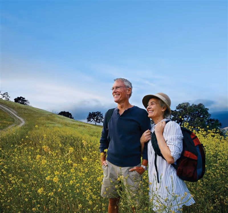 An older couple standing in a field of yellow wildflowers with a dirt path and trees in the background under a blue sky. The man is wearing a dark long-sleeve shirt and light shorts, and the woman is wearing a light striped shirt, a hat, and carrying a backpack. Both are smiling and looking into the distance.