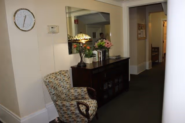 Interior hallway of a senior living facility with a patterned armchair, a dark wooden cabinet topped with a decorative lamp and flower arrangements, a large mirror on the wall, a clock, and a thermostat. The hallway extends into other rooms with beige walls and carpeted floor.