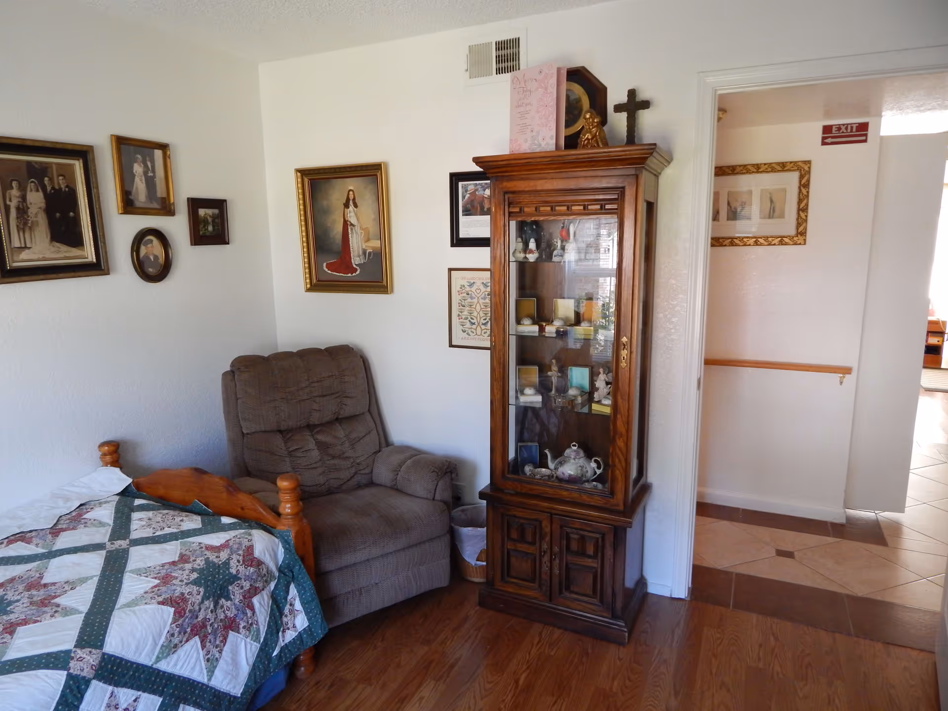 A cozy bedroom corner with a wooden bed covered by a quilted blanket, a cushioned recliner chair, and a tall wooden display cabinet filled with various decorative items. The walls are adorned with framed pictures and artwork, and there is a doorway leading to a hallway with tiled flooring and an exit sign above.